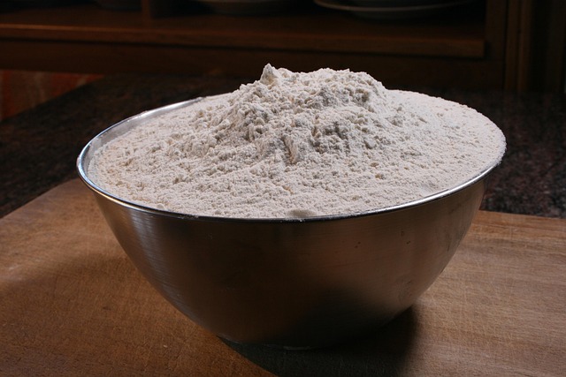 Flour being poured from its paper bag into a large, airtight glass storage container.