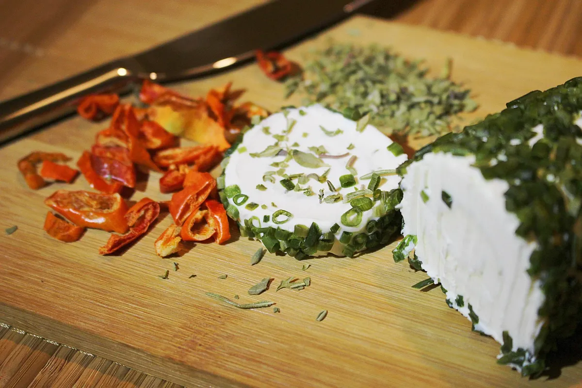 A round of soft white fresh cheese rolled in chopped chives, with dried herbs and dried tomato slices on a wooden cutting board
