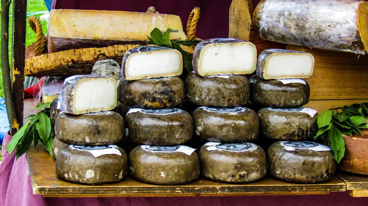 Stacks of aged goat cheese wheels with dark rinds, some cut open to show the white inside, on a wooden market stall